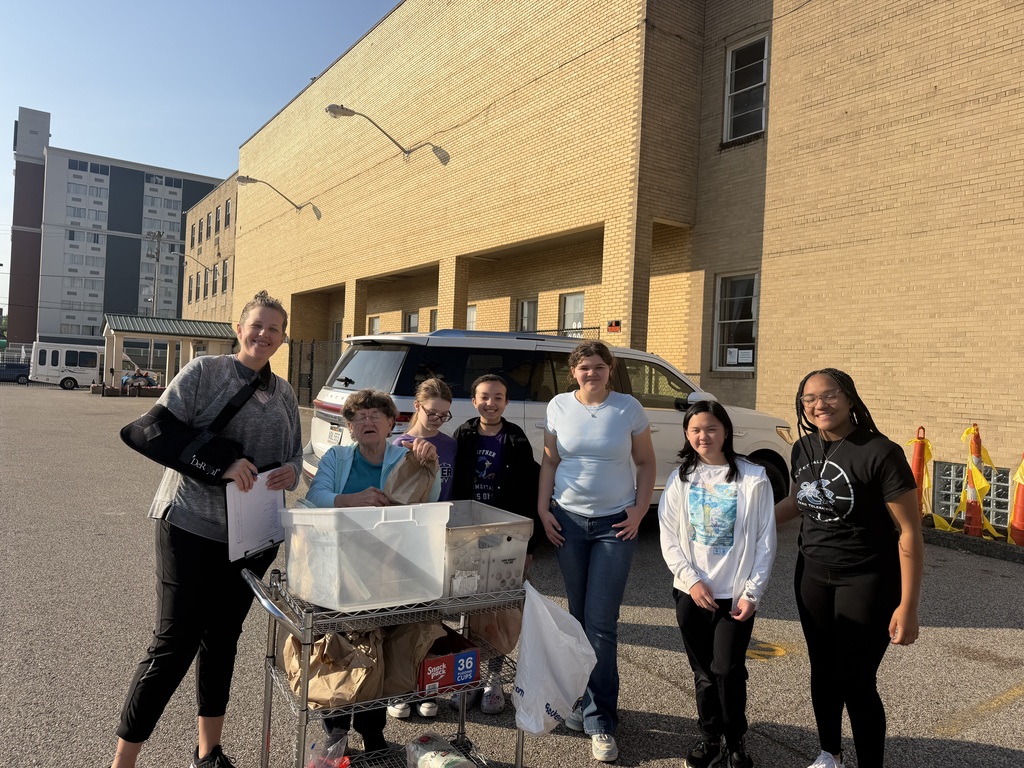 A group of students and their teacher smiling for a picture before starting their service project. There is a metal cart in front of the students that is holding different boxes and bags.