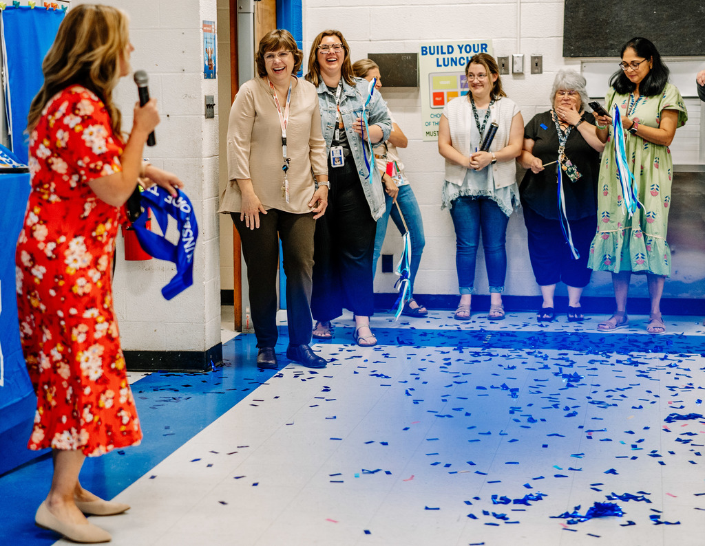 KCS Counselor of the Year, Emily Patterson standing in the doorway of the school's cafeteria and smiling as the school principal says kind things about Emily. There is blue confetti covering the floor.  