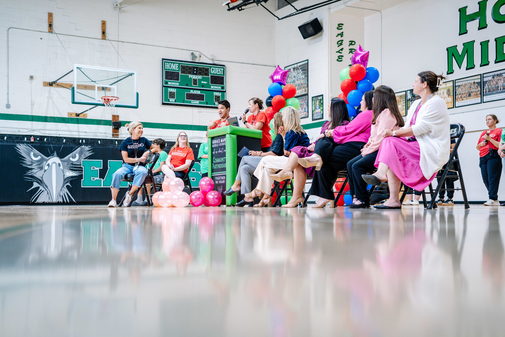 Horace Mann VP, Mrs. Harris, is speaking at the podium in the school's gymnasium to congratulate Ms. Tweedy. Ms. Tweedy is sitting on a chair in front of the podium. On the left side of the podium are the KCS assistant superintendents and superintendent.  