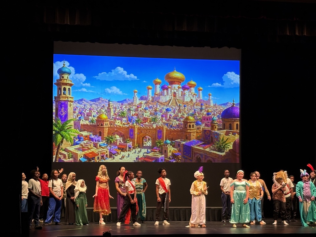 Students on stage during their performance of Aladdin. Students are dressed in early middle eastern outfits consistent with what the characters in the original Aladdin movie wore. Behind the students is a large screen that serves as the play's backdrop.  On the screen is a picture of Agrabah, the fictional city in which Aladdin takes place.  