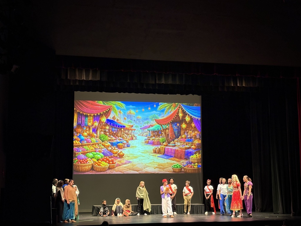 Students on stage during their performance of Aladdin. Students are dressed in early middle eastern outfits consistent with what the characters in the original Aladdin movie wore. Behind the students is a large screen that serves as the play's backdrop.  On the screen is a picture of Agrabah, the fictional city in which Aladdin takes place. 