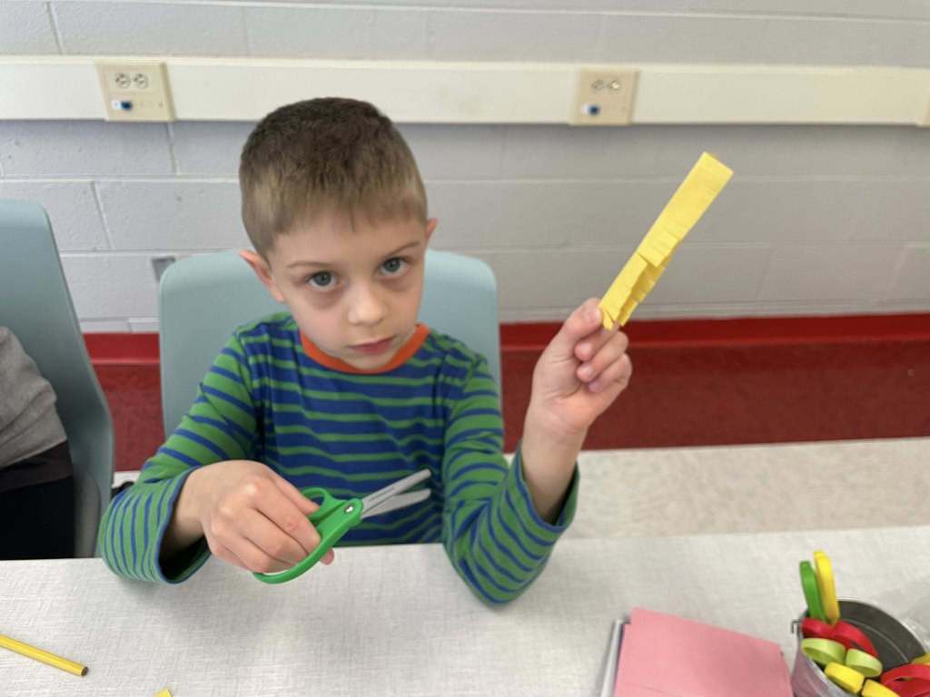 A student sitting at a table. The student is holding a yellow piece of paper in his left hand and green scissors in his other hand. The student has small lines in the yellow paper for his craft.