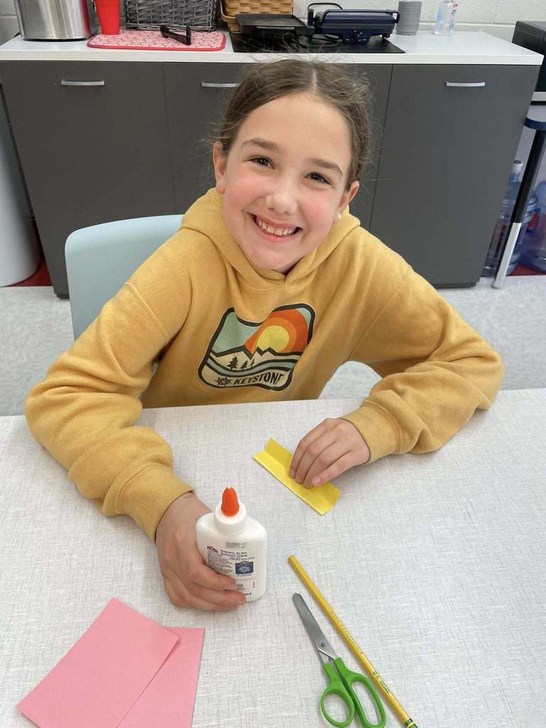 A student sitting at a table and smiling at the camera. The student is holding a bottle of glue in her right hand and a small piece of yellow paper in her left hand. The yellow paper has been folded in half. In front of the student are two pieces of pink paper, green scissor, and an unsharpened pencil.