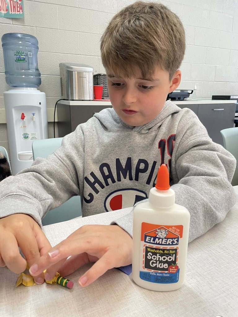 A student sitting at a table doing an art project. There is a bottle of glue by his left arm. He is holding a small piece of yellow paper to a pencil.