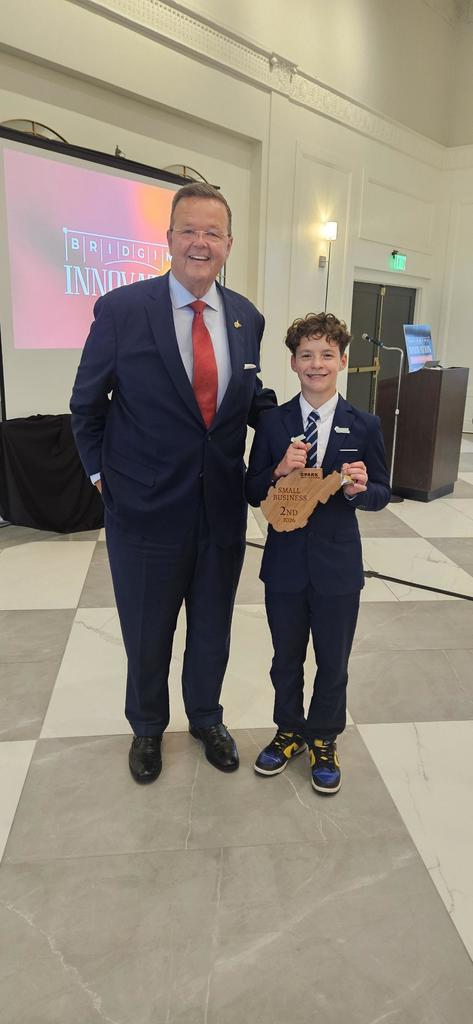 Parker, holding his 2nd place plaque. The plaque is a wooden cutout of the state of West Virginia. It says Small Business 2nd on it. Parker is standing with WV Secretary of State Kris Warner.