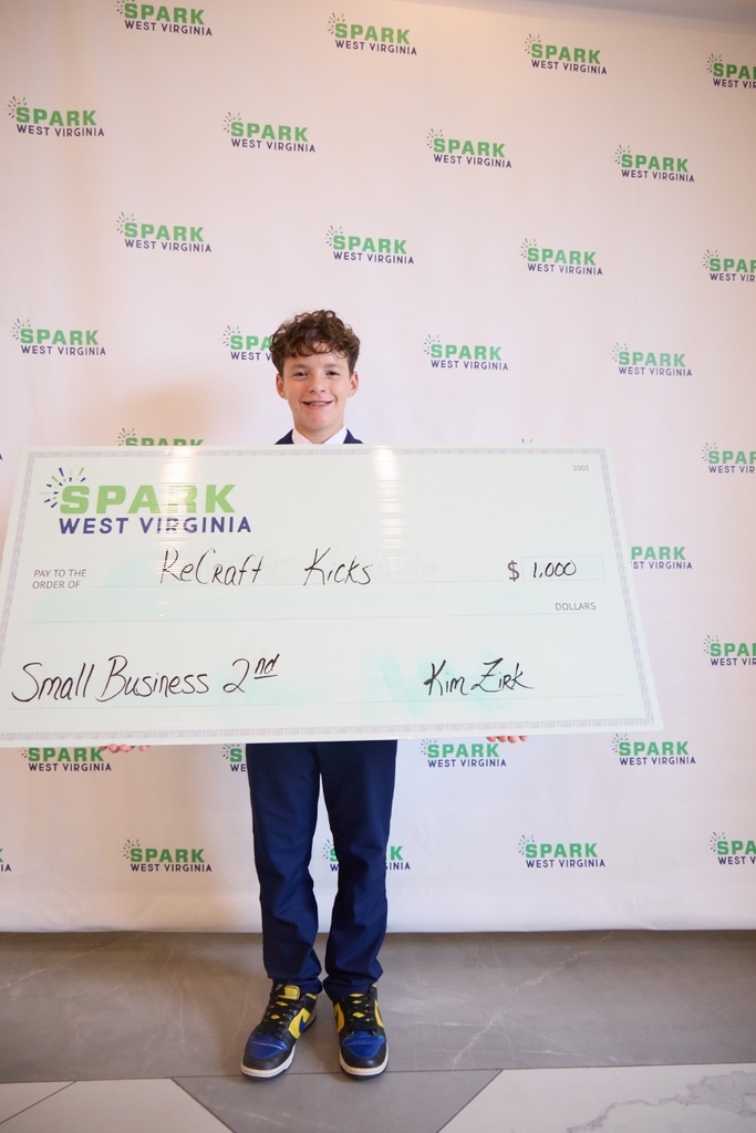 Parker is standing in front of a Spark West Virginia photo backdrop. He is holding an oversized prize check. The check is made out to his company ReCraft Kicks and is worth one-thousand dollars.