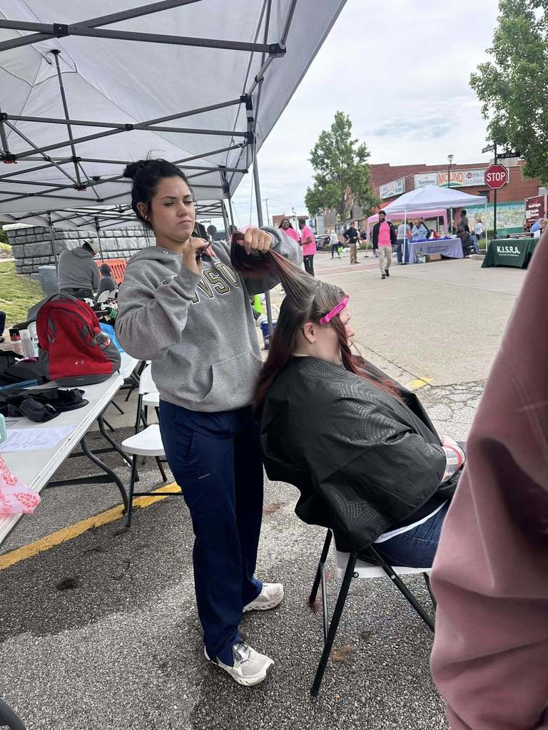 A Carver Beauty Academy Student is holding a woman's hair in her left and and scissors in the other hand. The woman receiving the haircut is seated and wearing a black barber cape. The student is standing behind the woman and getting ready to trim a small piece of hair with the scissors. The woman's hair is separated into different sections for the haircut with pink clips. 