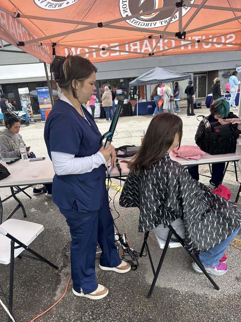 A Carver Beauty Academy Student standing behind a woman getting her hair styled. The student is holding a curling iron as she prepares to curl the woman's hair. The woman is seated in a chair and is wearing a barber cape. The cape is black with images of barber scissors. 