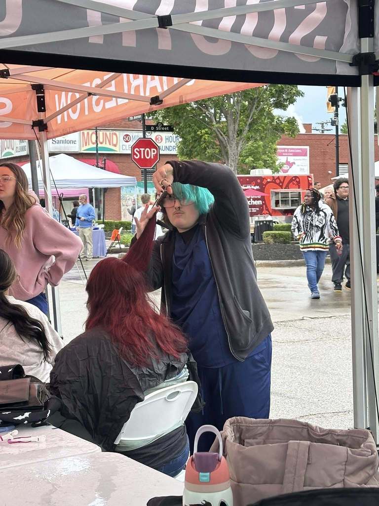 A Carver Beauty Academy is holding a piece of a woman's hair in one hand and scissors in the other hand and is getting ready to make a cut. The woman receiving the haircut is sitting down wearing a black barber cape, and the student is standing beside her. 