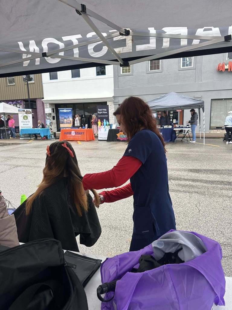A Carver Beauty Academy Student using scissors to cut a woman's hair. The woman is seated in a chair in front of the student. The woman has two hair clips holding her hair in place for the haircut.