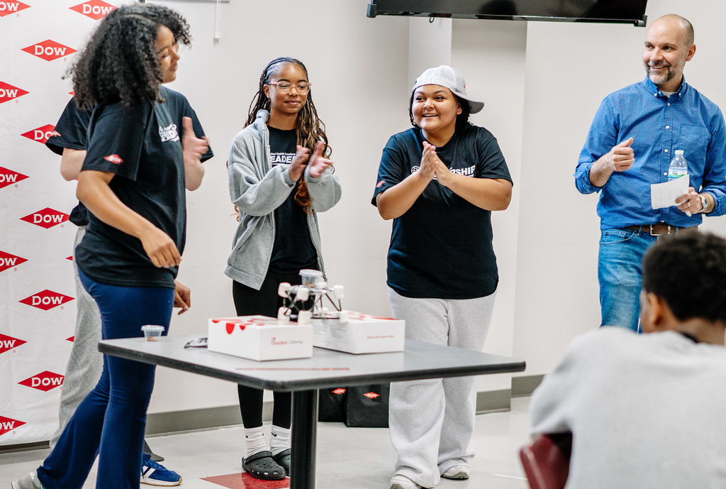 Four SCHS students are standing behind a table smiling and clapping. On the table in front of them is the model bridge they designed. The bridge is sitting on top of two Chic-Fil-A boxes that are slightly spread out. On top of the bridge is a weight that is testing the strength of the student's bridge. The bridge was able to hold the weight.