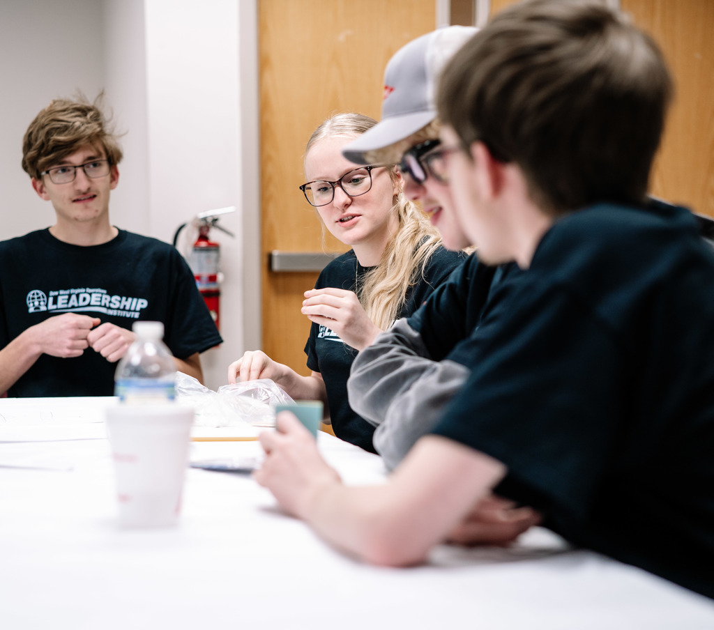 One student is sitting at the end of a table, and three others are sitting to his left on the longer side of the table. They are trying to figure out what their next step is to build their bridge. 