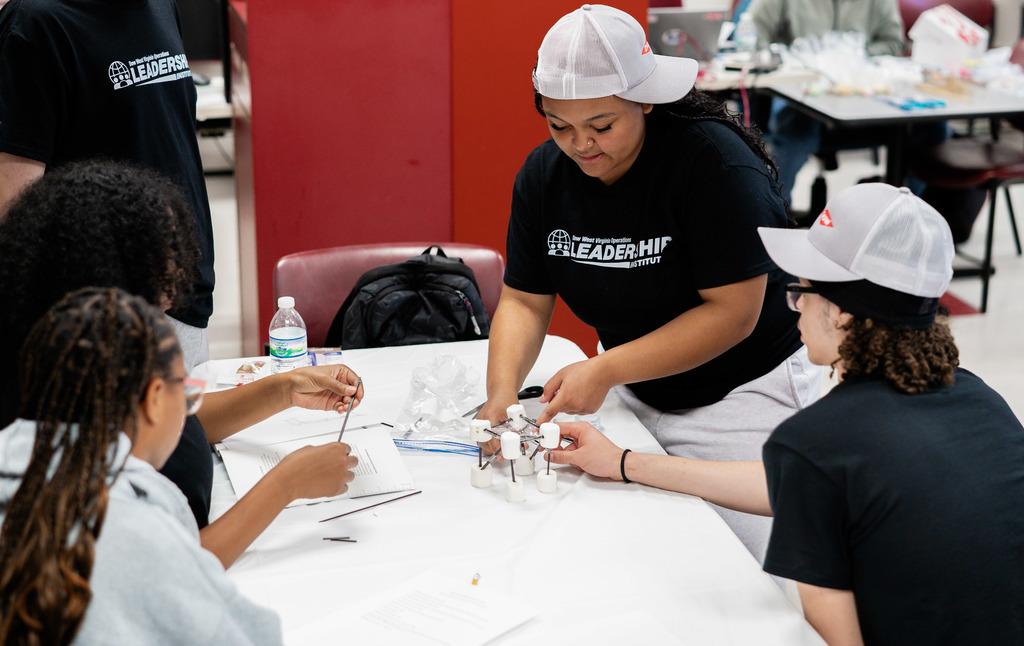 An SCHS student is standing at a table while she is working on her model bridge. Another student, to her left, is holding a different part of the bridge with his right hand. Two students are sitting across the table from them and looking on. 