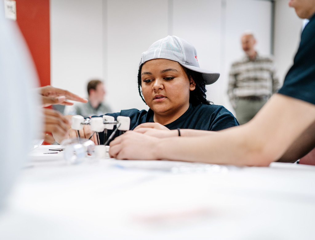 A student is squatting down behind a table while working on her model bridge. 