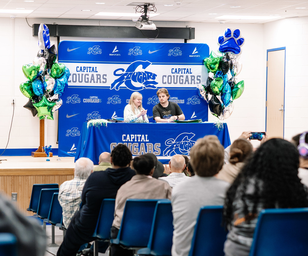A Marshall University representative sitting at a table on stage with a Capital High School student. The table has a Capital High School tablecloth on it. Behind the table is a Capital  High School Photo Wall. There are marshall and capital themed balloons on either side of the photo wall.