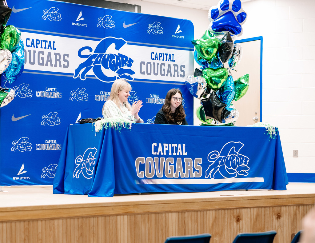 A Marshall University representative sitting at a table on stage with a Capital High School student. The table has a Capital High School tablecloth on it. Behind the table is a Capital  High School Photo Wall. There are marshall and capital themed balloons on either side of the photo wall.