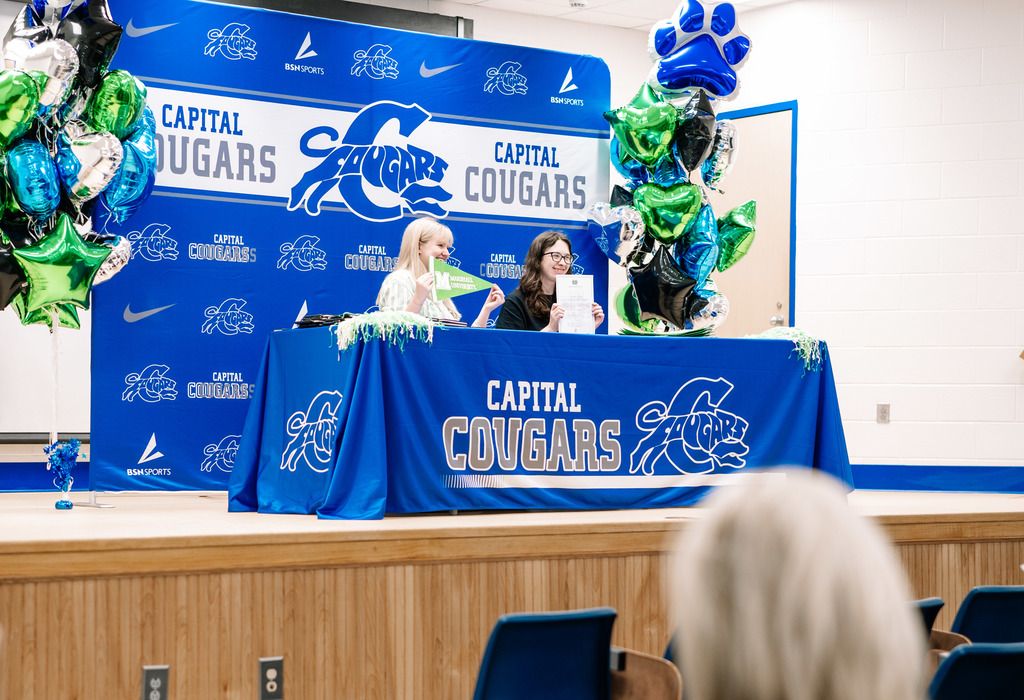A Marshall University representative sitting at a table on stage with a Capital High School student. The table has a Capital High School tablecloth on it. Behind the table is a Capital  High School Photo Wall. There are marshall and capital themed balloons on either side of the photo wall.