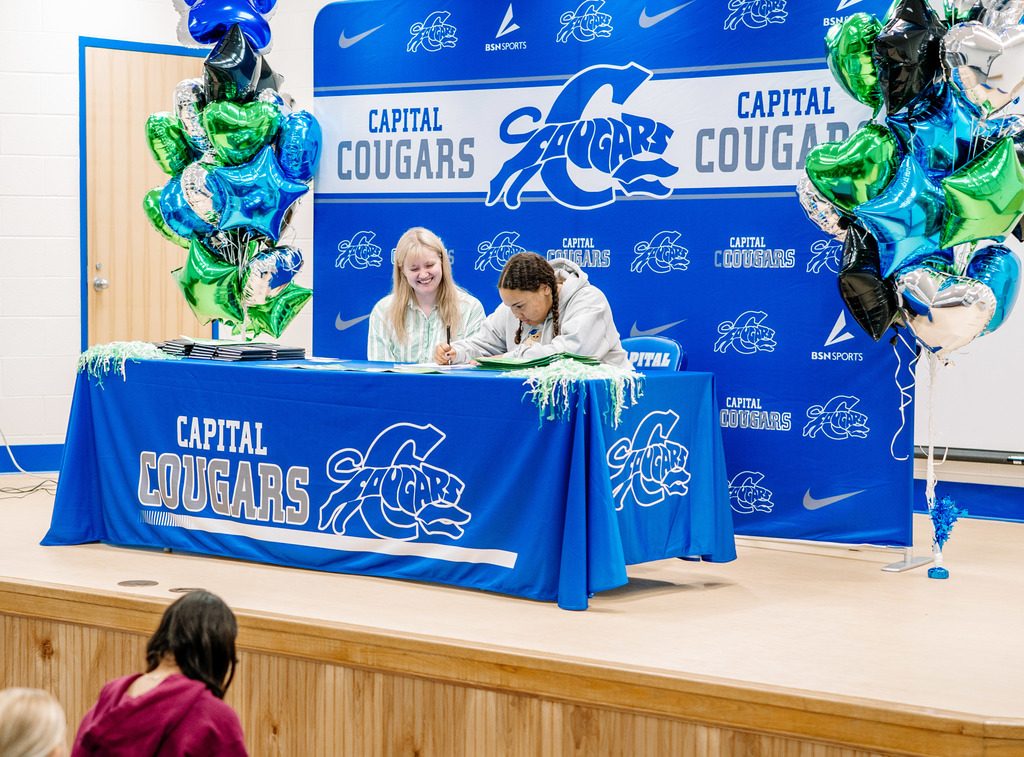 A Marshall University representative sitting at a table on stage with a Capital High School student. The table has a Capital High School tablecloth on it. Behind the table is a Capital  High School Photo Wall. There are marshall and capital themed balloons on either side of the photo wall.