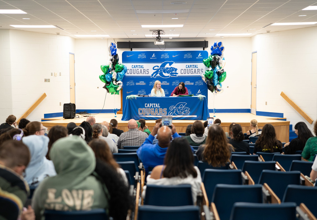 A Marshall University representative sitting at a table on stage with a Capital High School student. The table has a Capital High School tablecloth on it. Behind the table is a Capital  High School Photo Wall. There are marshall and capital themed balloons on either side of the photo wall.