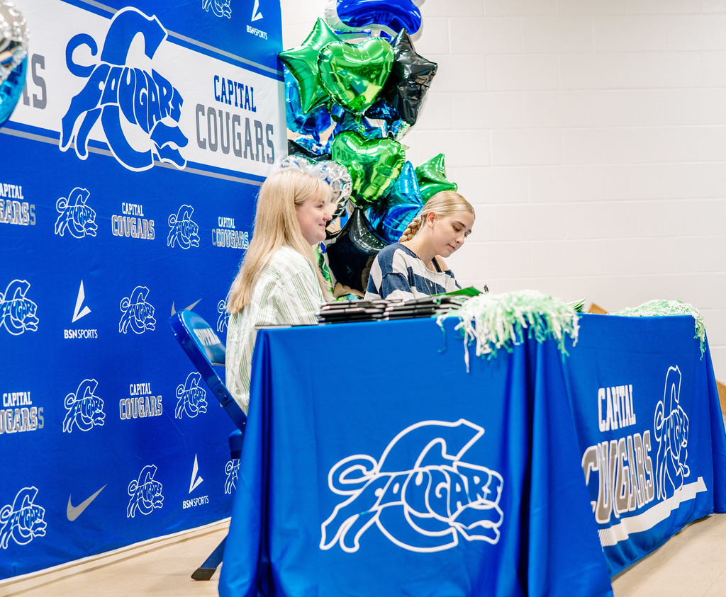 A Marshall University representative sitting at a table on stage with a Capital High School student. The table has a Capital High School tablecloth on it. Behind the table is a Capital  High School Photo Wall. There are marshall and capital themed balloons on either side of the photo wall.