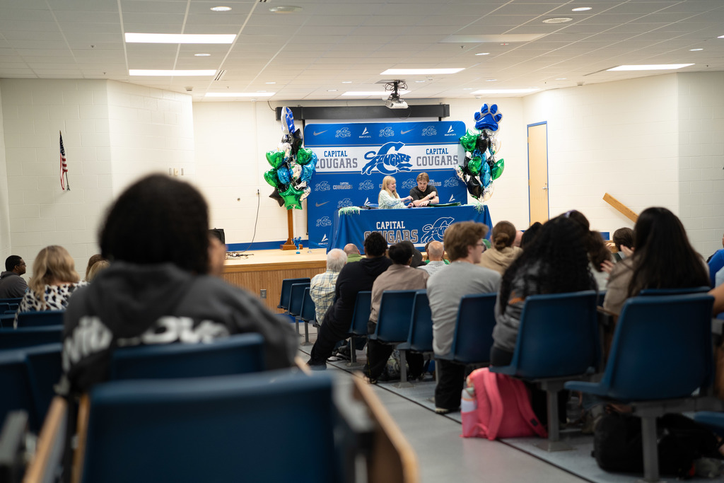 A Marshall University representative sitting at a table on stage with a Capital High School student. The table has a Capital High School tablecloth on it. Behind the table is a Capital  High School Photo Wall. There are marshall and capital themed balloons on either side of the photo wall.