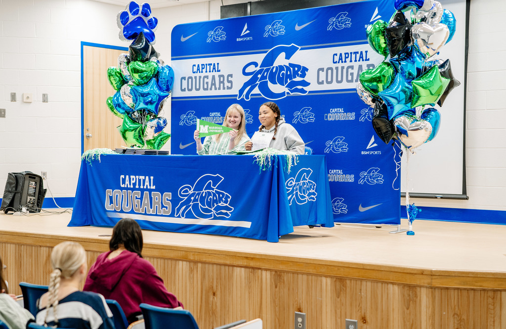 A Marshall University representative sitting at a table on stage with a Capital High School student. The table has a Capital High School tablecloth on it. Behind the table is a Capital  High School Photo Wall. There are marshall and capital themed balloons on either side of the photo wall.