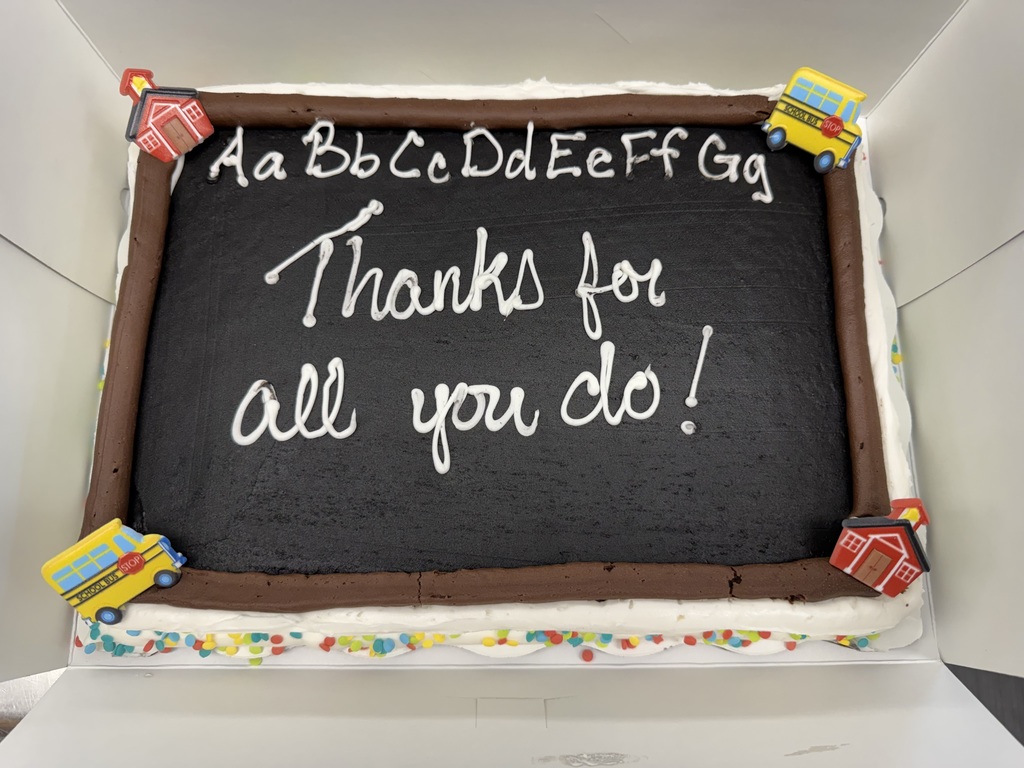 Chocolate cake designed as a chalkboard with white icing letters and "Thanks for all you do!" Bright bus and schoolhouse toppers add a cheerful touch.