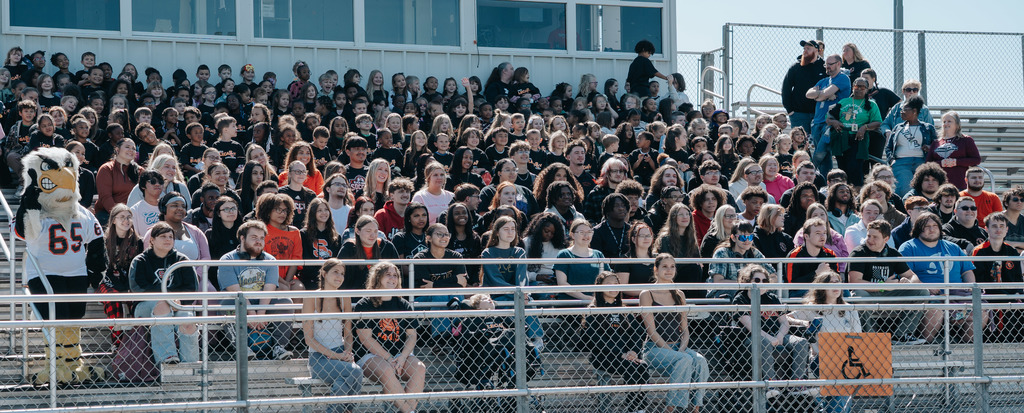 High school students and elementary students sitting in the bleachers for a large group photo. The seniors are in the first few rows and the elementary students are in the rows behind them. 