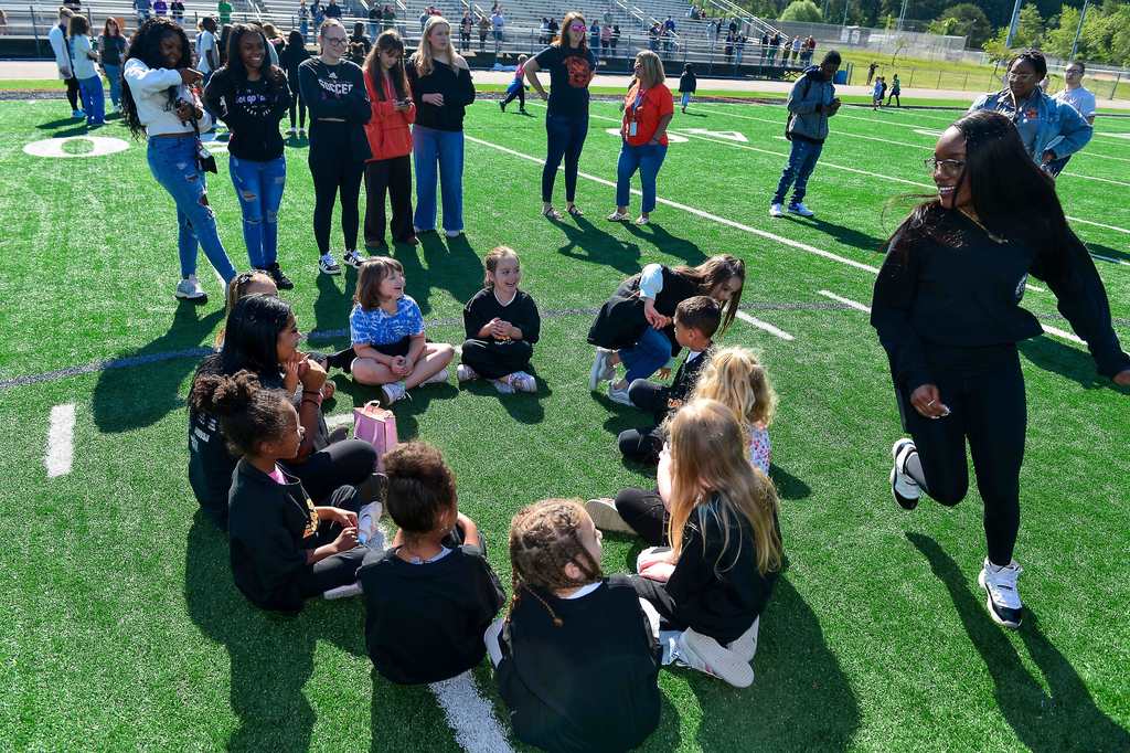 Young students sitting in a circle on the football field while a high school student is running around the circle. They are playing duck duck goose. 