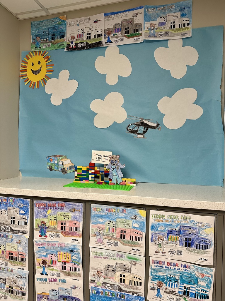 The Teddy Bear Fair coloring contest display created by Cedar Grove Elementary students. There are several coloring pages of a teddy bear and Women and Childrens Hospital hanging up, along with a hospital built with legos and a teddy bear cutout standing beside the hospital. The background of the display is blue construction paper with cloud cutouts, a sun, and a cutout of a helicopter that is colored black. 