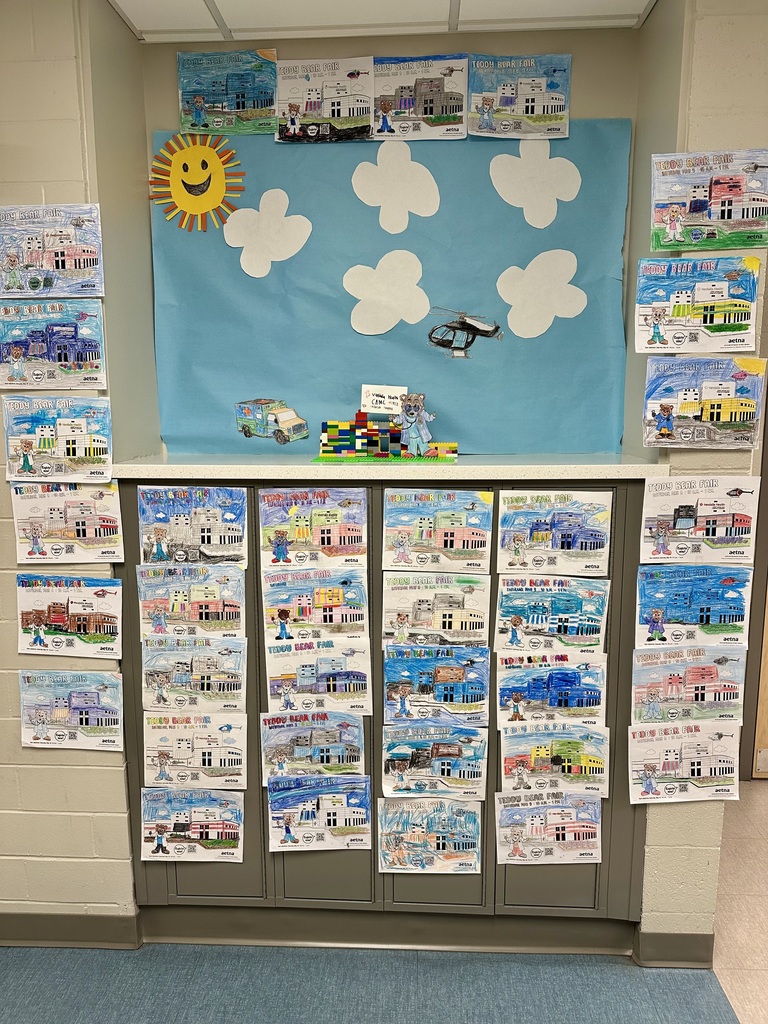 The Teddy Bear Fair coloring contest display created by Cedar Grove Elementary students. There are several coloring pages of a teddy bear and Women and Childrens Hospital hanging up, along with a hospital built with legos and a teddy bear cutout standing beside the hospital. The background of the display is blue construction paper with cloud cutouts, a sun, and a cutout of a helicopter that is colored black. 