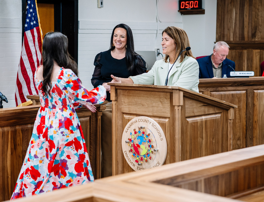 Dr. Potter standing behind a podium reaching out to shake a teachers hand as the teacher is walking up to receive her award. Asst. Superintendent, Amanda Mays, is standing to the side of Dr. Potter and is smiling. 