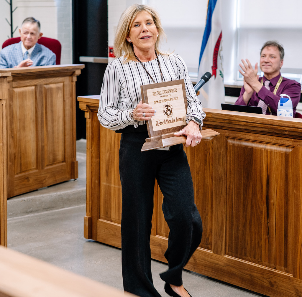 2026 KCS Principal of the Year, Shandon Tweedy, is walking back to her seat in the board room holding her principal of the year award. 