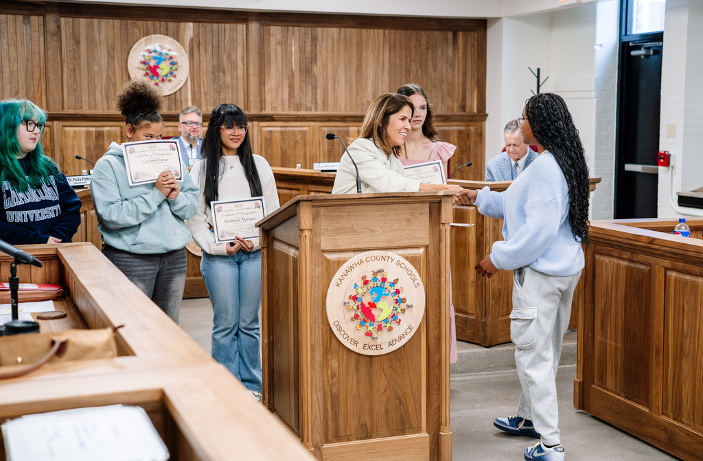 Dr. Potter standing behind a podium in the board room, shaking the hand of a student who won the 2026 Golden Horseshoe award. Four students who have already received their award are standing behind Dr. Potter. 