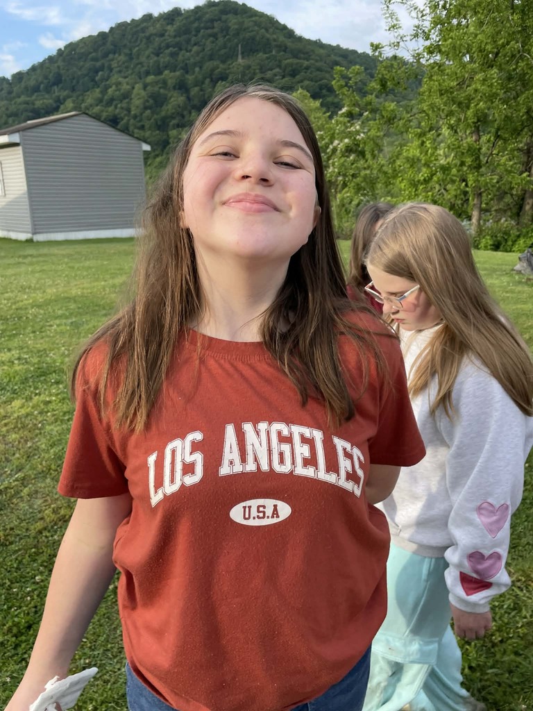 A girl smiles confidently, wearing a red "Los Angeles USA" t-shirt, with a scenic backdrop of a grassy field, trees, and a mountain. Another girl stands beside her.