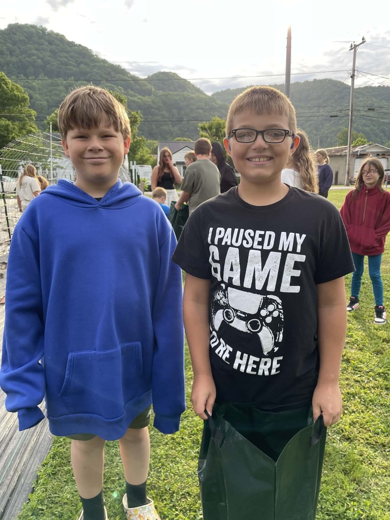 Two smiling boys stand on grass, one wearing a blue hoodie, the other a black "I Paused My Game" shirt. A scenic mountain backdrop and people are behind them.