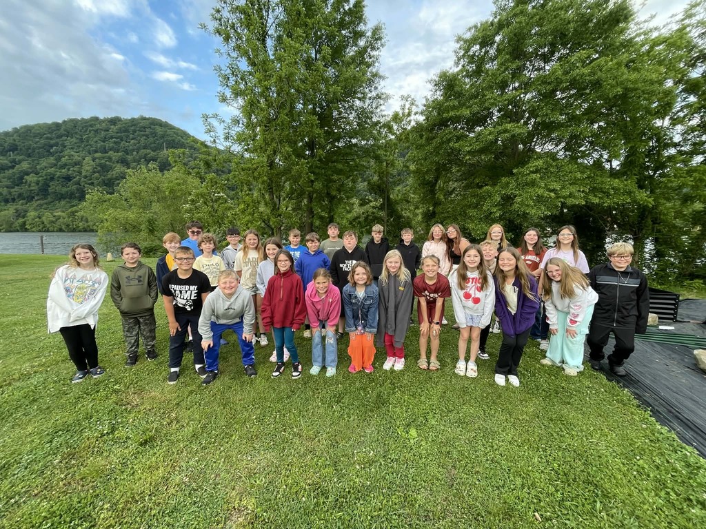 A group of children stands outdoors on a grassy area near a lake, surrounded by lush green trees and hills, under a partly cloudy sky.