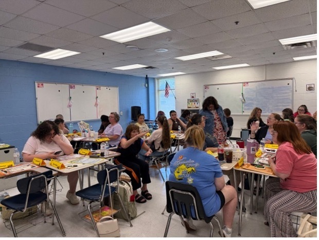 Teachers sitting at desks in a classroom. The desks have been pushed together in groups of three or four. They are working on an activity during a professional development session.