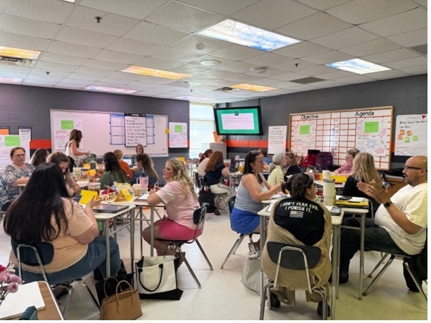 Teachers sitting at desks in a classroom. The desks have been pushed together in groups of three or four. They are working on an activity during a professional development session.