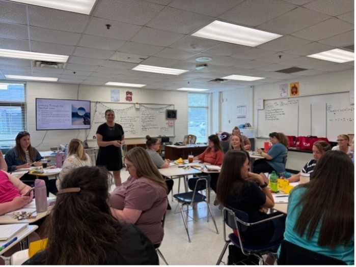 A classroom full of teachers. One woman is standing at the front and is leading the class.