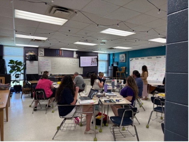 Teachers watching a video on a TV in a classroom while sitting in desks.