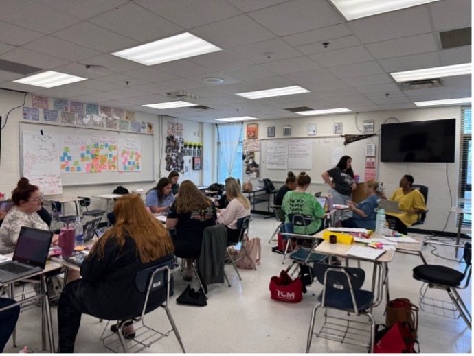 Teachers sitting at desks in a classroom. The desks have been pushed together in groups of three or four. They are working on an activity during a professional development session.