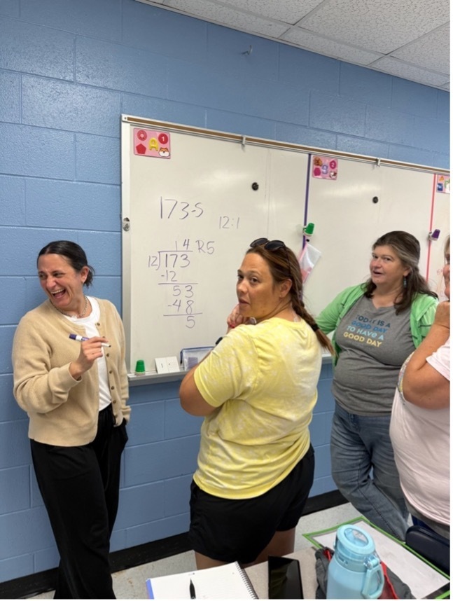 Four teachers are solving an equation on a white board in a classroom. They are looking at someone off camera and laughing.