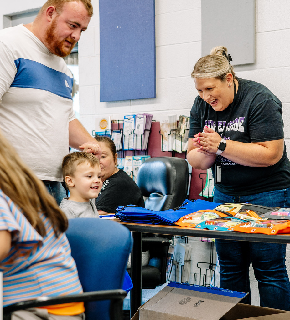 A young student and his father at the registration table for the open house. The student is walking in front of his dad while holding his hand. A KCS employee is smiling and clapping as the student walks in.
