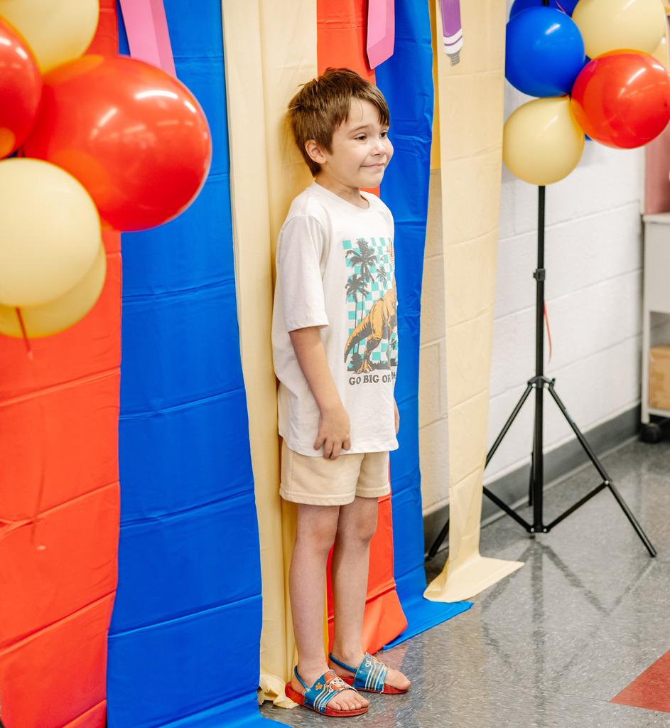 A young student standing in front of a red, blue, and yellow photo wall and smiling. There are also balloons on either side of the student. 