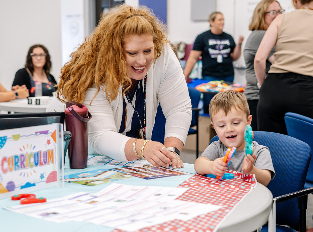 A KCS employee and a young student playing with a toy at the curriculum table at the open house. The student has a toy and each hand and the employee is smiling watching him play.