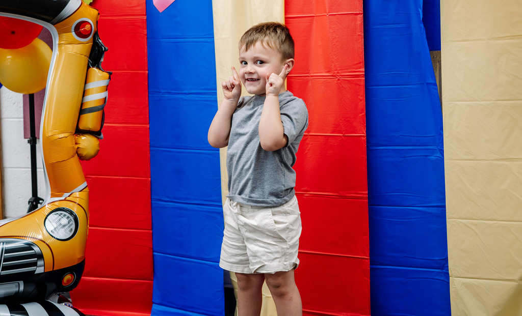 A young student standing in front of a red, yellow, and blue photo wall. He is smiling and is pointing both of his index fingers towards the ceiling. 
