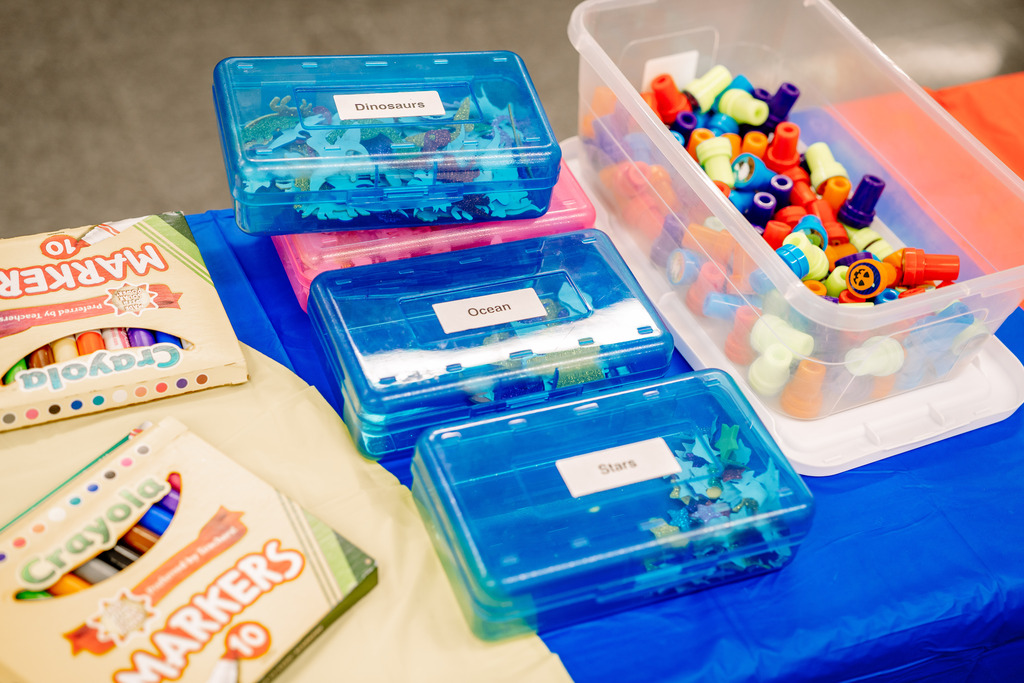 Two boxes of markers sitting on an activity table. Three blue pencil boxes filled with dinosaurs action figures, ocean toys, and star stickers. There is also a clear plastic bin with stamps inside them.