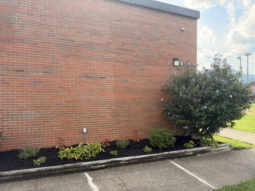 Red brick building wall with landscaped flower bed, flowering shrub, and empty parking spaces under a bright, partly cloudy sky.