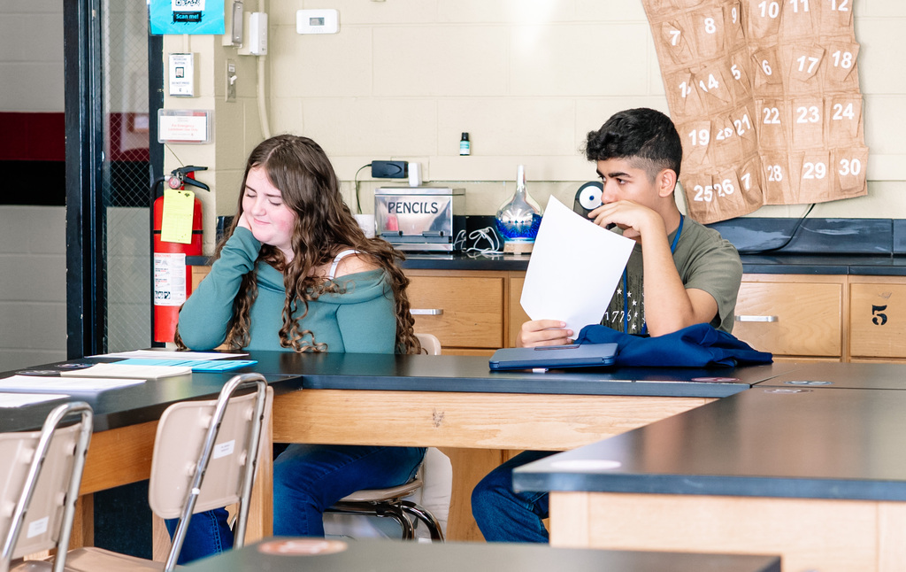 Two students sitting at a classroom table. One his holding his paper over his mouth and the other is smirking while looking down at a paper in front of her.
